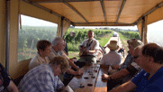 Blick in den Planwagen, die Gruppe zu beiden Seiten, in der Mitte der Tisch mit gefüllten Gläsern, Achim mit Gitarre  auf dem Kutschbock neben der Kutscherin.