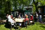 die ganze Gruppe sitzt um einen runden Tisch im Garten, im Hintergrund das Haus und die vorne offene überdeckte Terrasse.