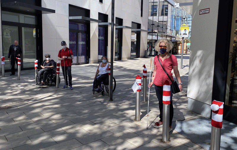 Die Gruppe, 2 Rollstuhlfahrer, zwei mit Blindenstock, darunter vprne Silvia schäfer, an 9 Pollern mit rot-weiß gestreiften Mützen