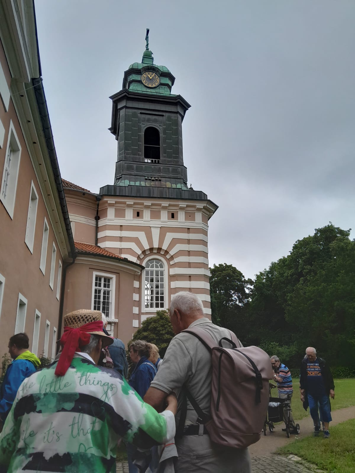 Einige der Reisenden stehen vor einem Glockenturm. Die Uhr zeigt 11:05 Uhr. Auf der rechten Seite schliet sich ein Park an das Gebude an.